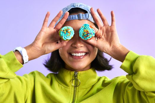 Spread positivity around like sprinkles. Studio shot of a young woman holding cupcakes over her eyes against a purple background.