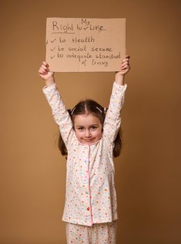 Adorable Caucasian preschool girl holding cardboard poster promoting children's rights to an adequate standard of living, social secure and health care, isolated on a beige background with copy space