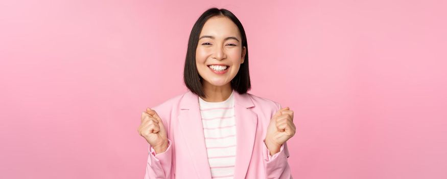 Enthusiastic saleswoman, asian corporate woman say yes, achieve goal and celebrating, triumphing, looking with rejoice and smiling, standing over pink background