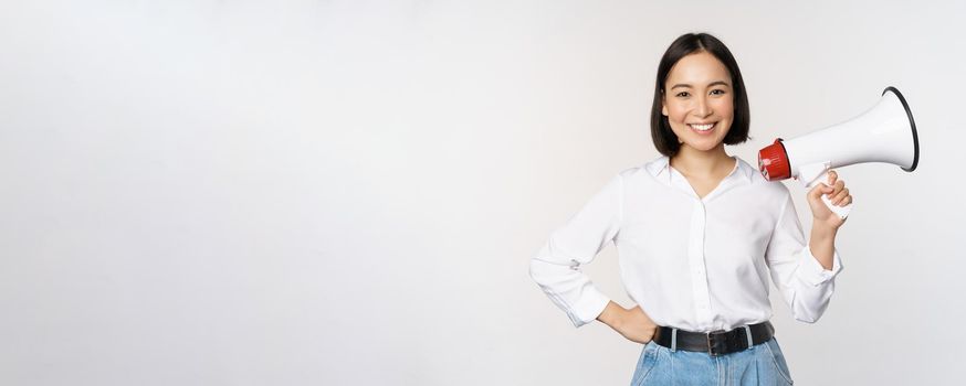 Smiling young asian woman posing with megaphone, concept of news, announcement and information, standing over white background.