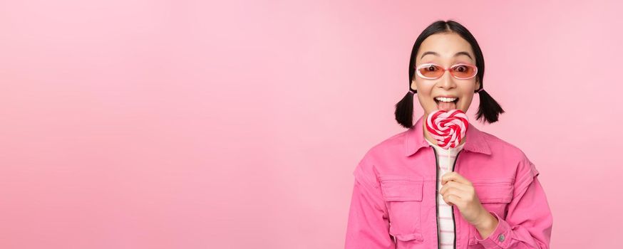 Stylish korean girl licking lolipop, eating candy and smiling, standing in sunglasses against pink background