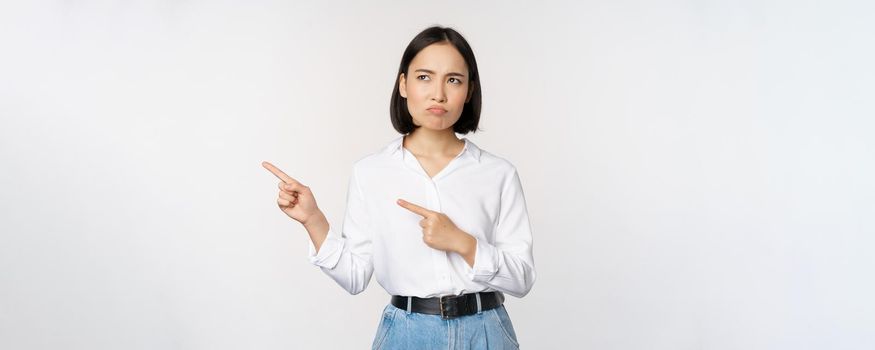 Disappointed, sad young asian woman pointing and looking left with upset, sulking face expression, standing over white background
