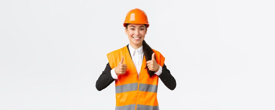 Upbeat pleased asian female architect giving permission, proud with result, standing in safety helmet and reflective jacket, showing thumbs-up in approval, guarantee best construction quality