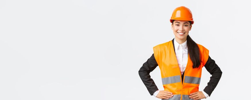 Successful young female asian businesswoman, architect in safety helmet, reflective jacket, standing confident and determined, smiling assured, building houses, posing over white background