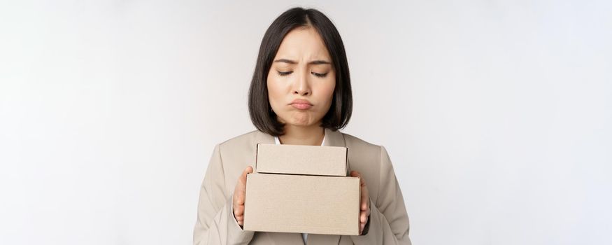 Portrait of asian saleswoman, female entrepreneur holding boxes and looking sad, disappointed, standing over white background