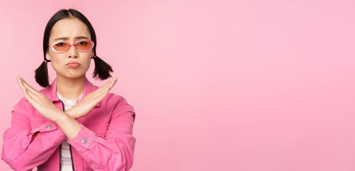 Portrait of korean girl in stylish sunglasses, sulking disappointed, showing stop, rejection gesture, cross sign, standing over pink background
