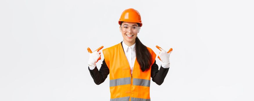Smiling confident asian female chief construction engineer in safety helmet, gloves and glasses pointing at herself, notify to follow safe protocol and wearing special clothing, white background