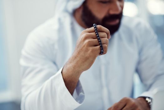 Seeking divine intervention. Cropped shot of a young businessman dressed in Islamic traditional clothing holding prayer beads while working in his office.