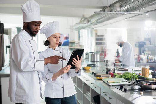 Food industry workers with handheld touchscreen device preparing ingredients for evening dinner service at restaurant. Gourmet cuisine experts with modern tablet researching ingredients on internet.