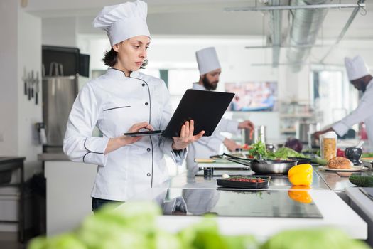 Sous chef with modern laptop looking for gourmet dish recipe while standing in restaurant professional kitchen.