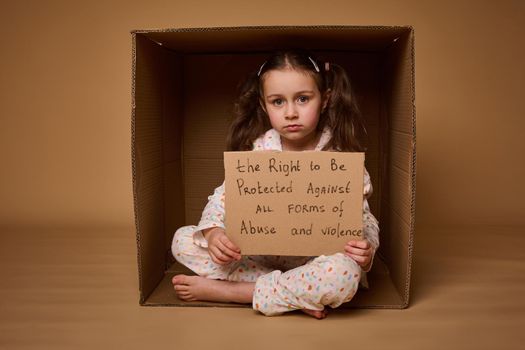 Confident baby girl holding a poster with a message to be protected from abuse and violence, sitting inside a cardboard box. Social advertisement for International Children day. 1st July
