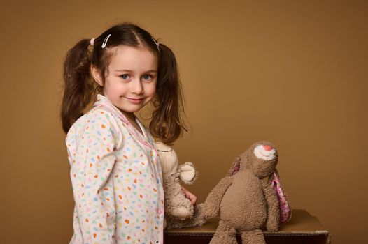 Close-up cute baby girl with two ponytails holding her plush toys and cutely smiles confidently looking at camera, isolated over beige background with copy ad space