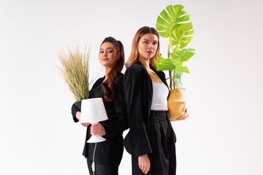 Two young business woman dressed black suit smile holding houseplant and lamp standing isolated on white background. Moving concept. Relocation new office with positive.