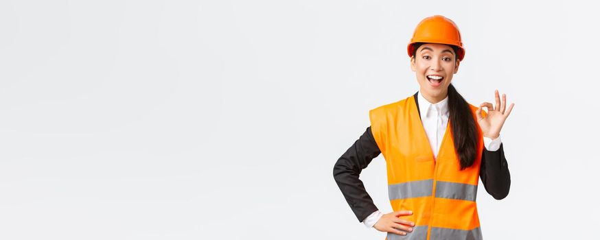 Satisfied happy asian female engineer, construction manager in safety helmet and reflective jacket showing okay gesture pleased, guarantee building quality, ensure everything excellent