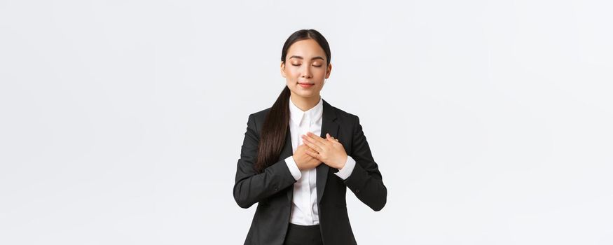 Happy dreamy asian female entrepreneur in suit close eyes and holding hands on heart, remember lovely memory, thinking about something pleasant over white background