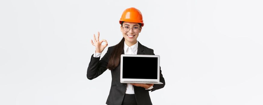 Smiling professional female architect, construction manager at factory showing graph, positive diagram, make okay gesture in approval and holding laptop facing screen, wear safety helmet