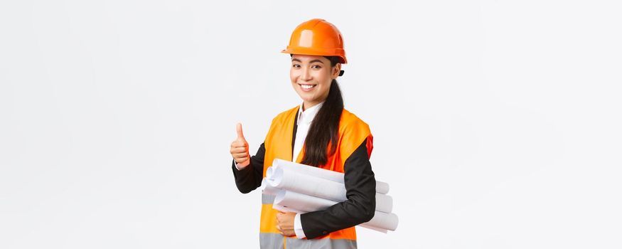 Confident successful female asian construction manager, architect in safety helmet and jacket, showing thumb-up and carry blueprints of building project, guarantee quality, white background