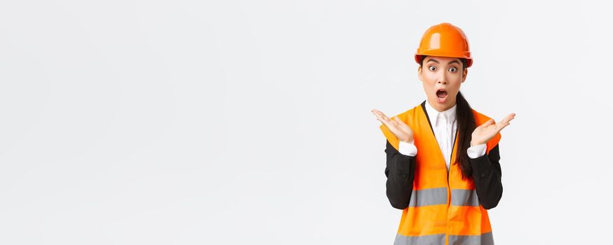 Surprised and shocked asian female engineer in reflective jacket, safety helmet, react to unexpected news, raising hands up and clap impressed, visit construction area, pleased with great work