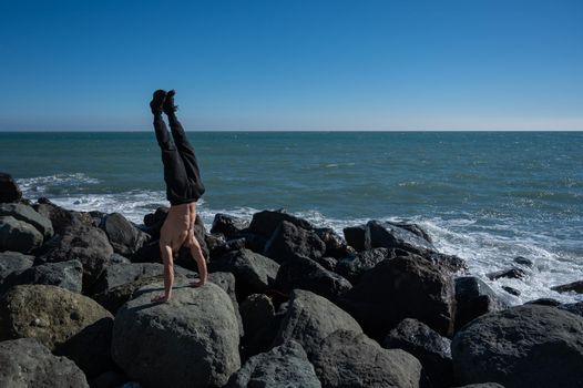 Shirtless man doing handstand on rocks by the sea.