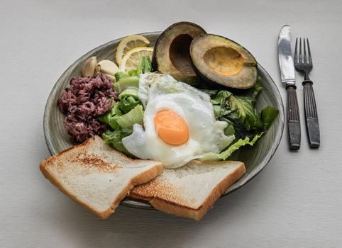 Breakfast with Fried Egg to put it on top of Fresh vegetables, Rice berry, Breads, and Garlic with Avocado cut in half and lemon sliced on Ceramic plate. 