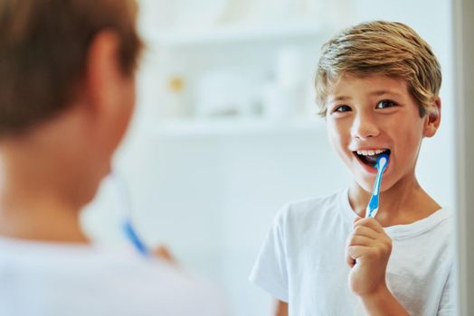 Shot of a cheerful young boy looking at his reflection in a mirror while brushing his teeth in the bathroom at home during the day.
