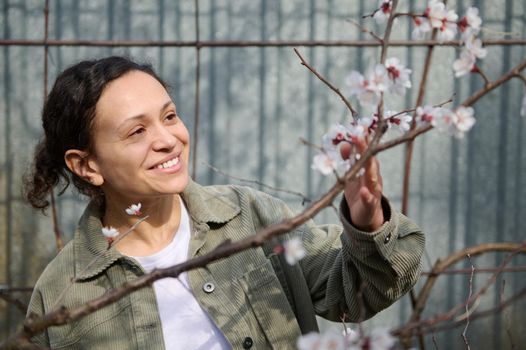 Pleasant woman smiles, enjoying sunny early spring day standing near blossoming fruit tree