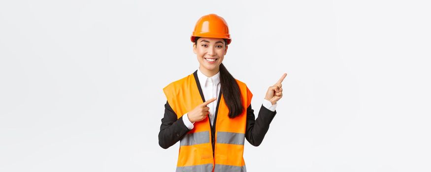 Building, construction and industrial concept. Smiling female asian engineer in safety helmet and reflective clothing showing object, pointing fingers right, saleswoman introduce estate to client