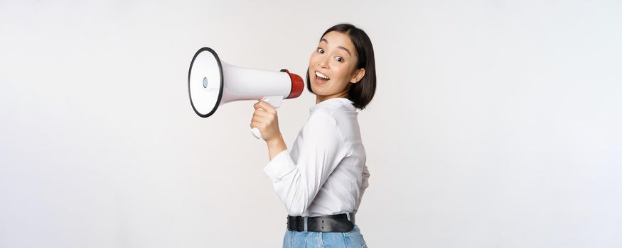 Beautiful young asian woman talking in megaphone, screams in speakerphone and smiling, making announcement, shout out information, standing over white background