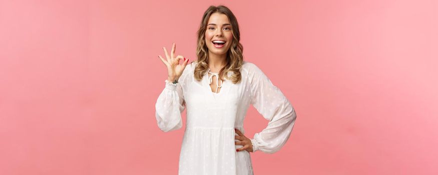 Portrait of carefree good-looking blond girl, wear white dress, guarantee you will like this special spring offer, show okay sign, agree or accept, smiling and nod in approval, pink background