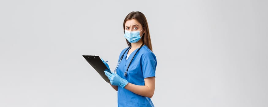 Covid-19, preventing virus, health, healthcare workers and quarantine concept. Suspicious and skeptical female nurse, doctor in blue scrubs fill-in patient forms on clipboard turn camera puzzled