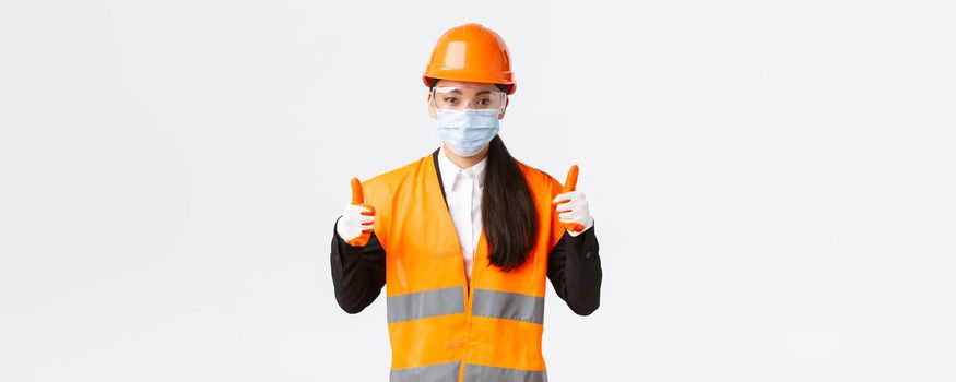 Covid-19 safety protocol at enterpise, construction and preventing virus concept. Confident female asian industrial worker, engineer in face mask and helmet showing thumbs-up, all good