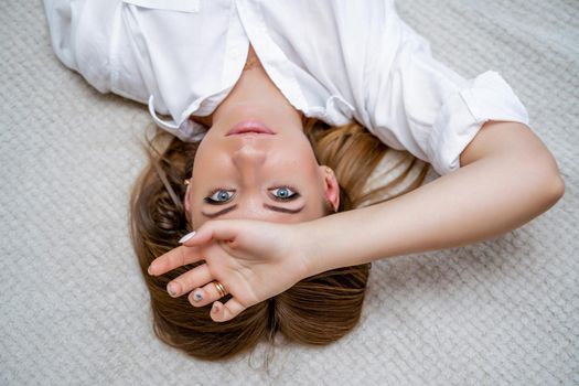 The woman lies on the bed on her back, top view. She looks straight ahead, wearing a white shirt