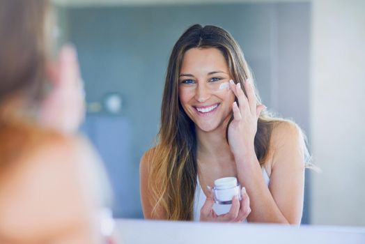 I moisturize daily for soft, smooth skin. Portrait of a beautiful young woman applying moisturizer to her face while looking in the mirror.