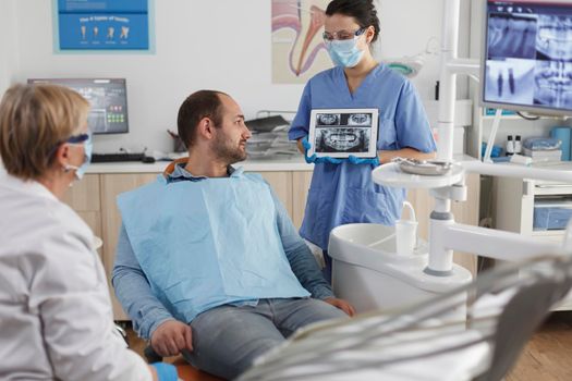 Dentist assistant with face mask holding tabet computer explaining teeth radiography to sick patient during dentistry consultation in stomatological office room. Team working at caries treatment