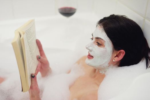 Treating herself to some time in the tub. Cropped shot of a young woman relaxing in the bathtub with a book and glass of wine.