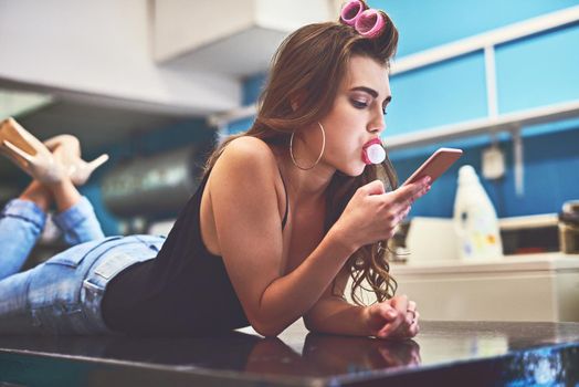 Nothing will get in the way of her doing what she wants. Shot of an attractive young woman lying on top of a counter while browsing on a cellphone to pass time in a laundry room.