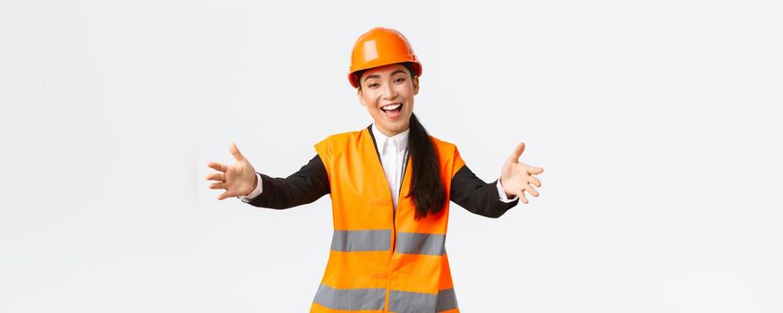 Friendly smiling asian female construction manager, engineer in safety helmet and reflective jacket, extending hands for greeting, welcome business partners, standing white background