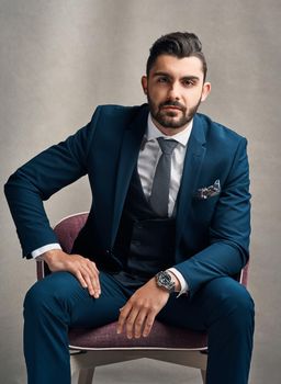 That CEO kinda style. Studio portrait of a stylishly dressed young businessman sitting on a chair against a grey background.