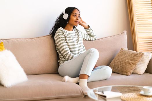 African-american female listening to audiobook in earphones while chilling on sofa in living room