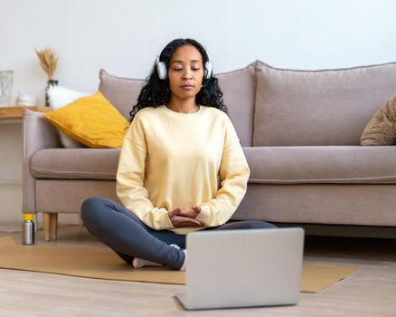African-american female listening to music in headphones while meditating with laptop at home