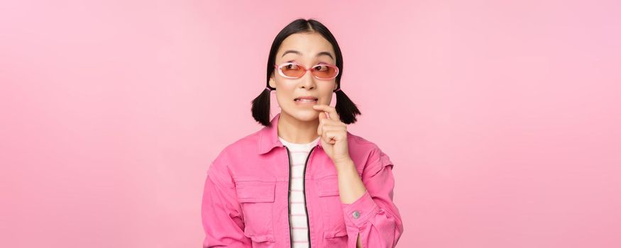 Stylish asian girl in sunglasses, looks thoughtful or intrigued, wants to try smth out, stands over pink background, pondering