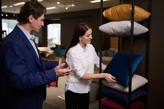 Retail assistant and customer near stand with sofa pillows in furniture store showroom