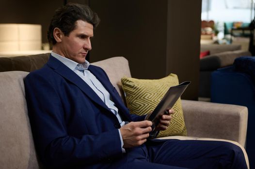 Confident mature man sitting on a sofa in showroom and leafing a catalog with upholstered furniture