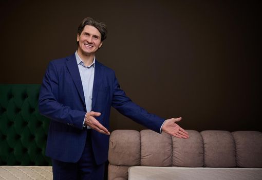 Handsome man sales manager consultant pointing at an upholstered bed and orthopedic mattress in the showroom of furniture store