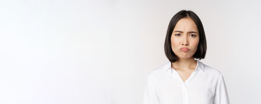 Sad and gloomy young asian woman grimacing, frowning upset, making pouting face, white background