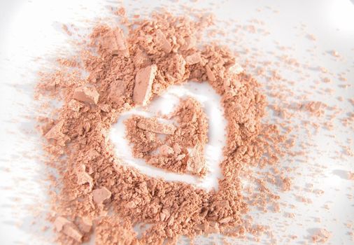 Scattered crumbs of face powder or eye shadow in the shape of a heart on a white background, makeup concept