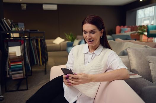 Interior designer- charming elegant brunette European woman in casual attire sitting on a stylish modern pink sofa and typing text on her mobile phone
