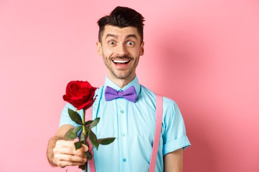 Valentines day and romance concept. Happy smiling man giving you red rose on romantic date, standing on pink background in bow-tie and shirt.