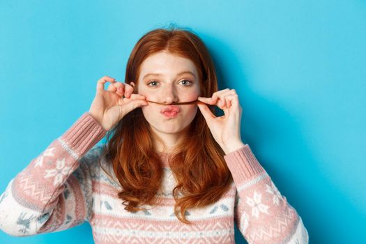 Close-up of silly and funny redhead girl making moustache with hair strand and puckered lips, grimacing against blue background