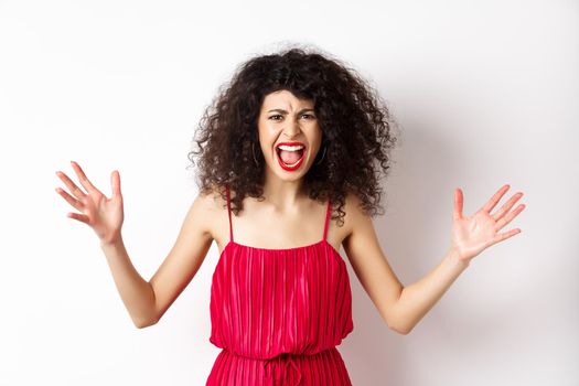 Angry young woman with curly hair, wearing red dress, screaming and having an argument, looking with hatred and anger, standing over white background